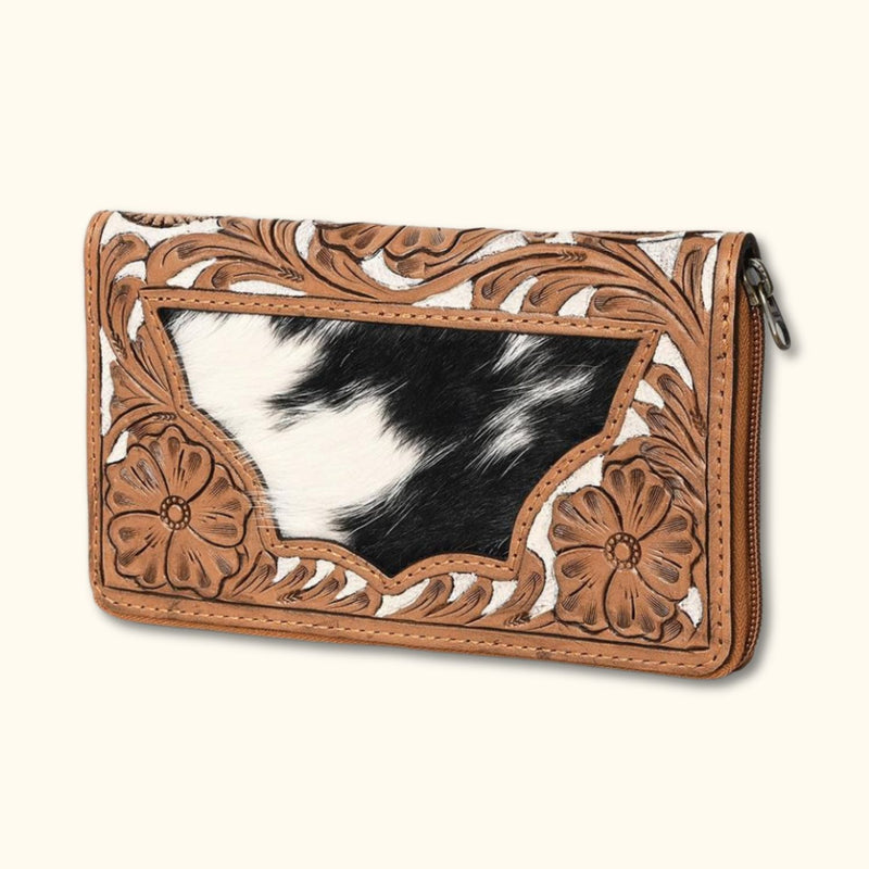 Angled view of a Western-style hair-on cowhide zip clutch with floral tooling and cowhide accent.