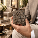 The Compact Crocodile Wallet in taupe green held in hand with a tailored neutral suit in a refined luxury setting