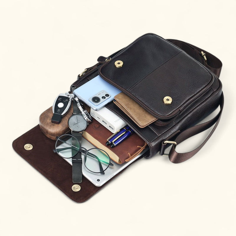 The Dustwalker full-grain cow leather crossbody iPad bag opened to show capacity with smartphone, glasses, watch, wallet, pens, and tablet inside.