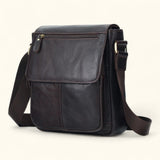 Angled side view of The Dustwalker full-grain cow leather crossbody iPad bag in coffee brown with adjustable strap and front flap design.