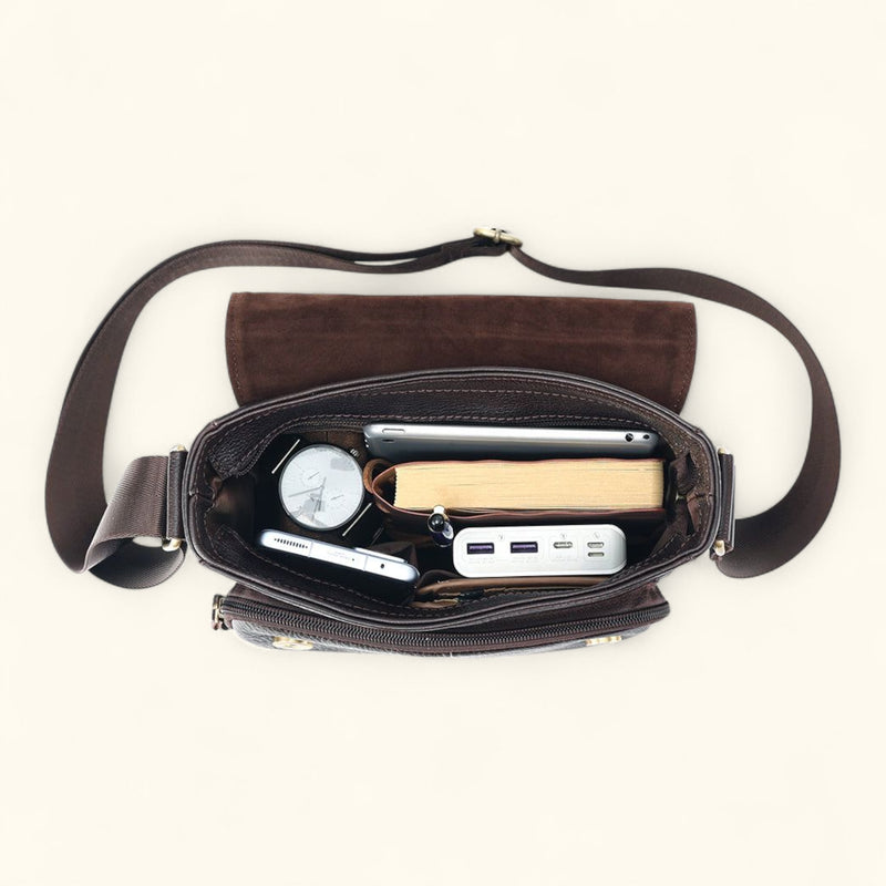 Top view of The Dustwalker full-grain cow leather crossbody iPad bag packed with watch, smartphone, power bank, notebook, and accessories inside.