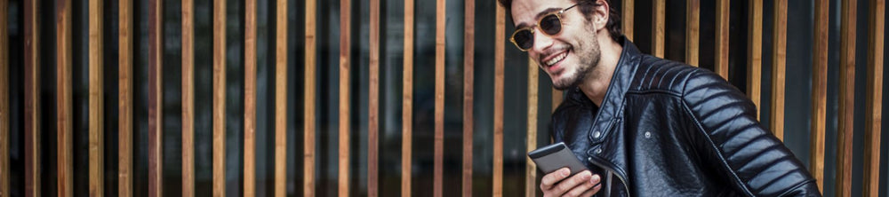 A stylish man wearing sunglasses and a black leather jacket, smiling and holding a smartphone while leaning against a wooden fence, representing the leather jacket collection.