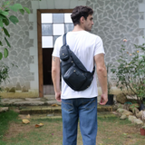 Man wearing a dark blue leather sling bag, standing outside with his back to the camera, dressed in a white t-shirt and blue jeans.
