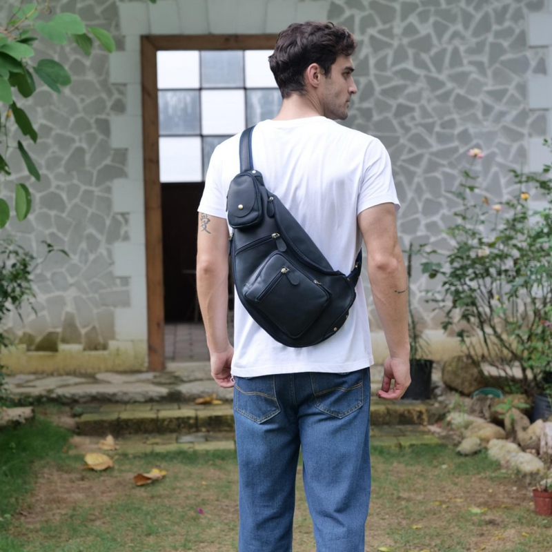 Man wearing a dark blue leather sling bag, standing outside with his back to the camera, dressed in a white t-shirt and blue jeans.
