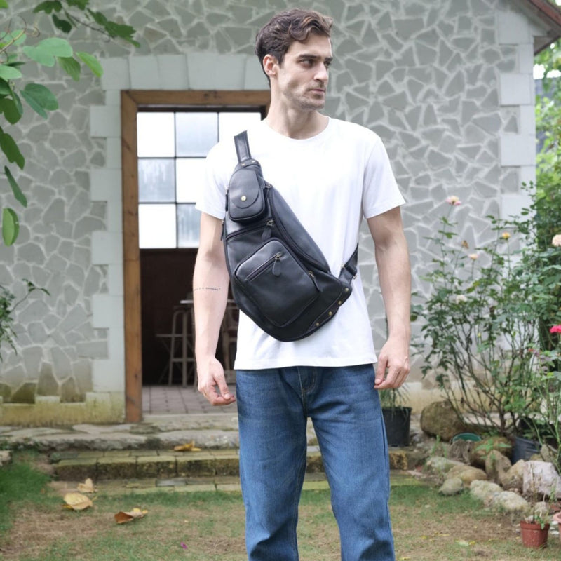 Man wearing a dark blue leather sling bag, standing outside facing the camera, dressed in a white t-shirt and blue jeans.