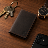 The Rancher's Legacy brown cowboy leather cardholder beside keys, a wristwatch, and a phone on a refined wooden surface.