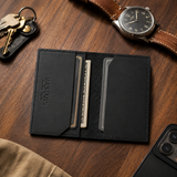 The Rancher's Legacy black Western leather cardholder open with cards and folded cash beside keys, a watch, and a phone on a wooden table.