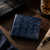 The Monarch Crocodile Bifold Wallet in Deep Midnight Blue displayed in a luxury lifestyle setting on a walnut desk alongside a fountain pen, whiskey glass, and premium accessories, showcasing the elegance of genuine crocodile leather craftsmanship.