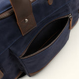 Close-up of The Weekender navy waxed canvas and leather duffel featuring an open front zippered pocket, reinforced leather handle attachment, and metal hardware.