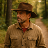 A man in his forties wearing a The Trailsman brown suede Western hat while walking through a forest trail, dressed in an outdoor work shirt with warm natural lighting.