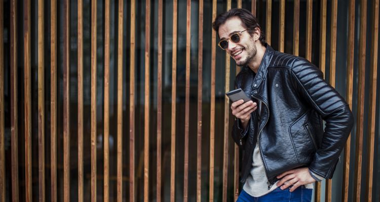 A fashionable man in a black leather jacket and sunglasses, casually leaning against a wooden fence, exemplifying modern urban style for the leather jacket collection.